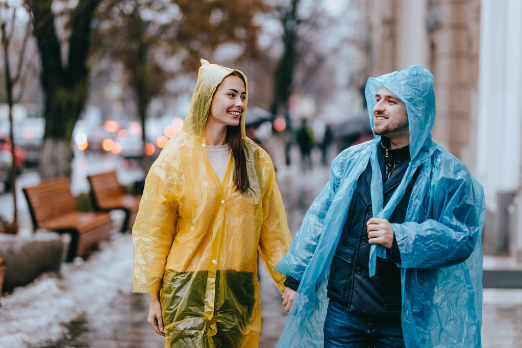 Sarı ve mavi yağmurluklar sevgi dolu adam ve kız yağmurda sokakta yürümek - Fotoğraf, Görsel
