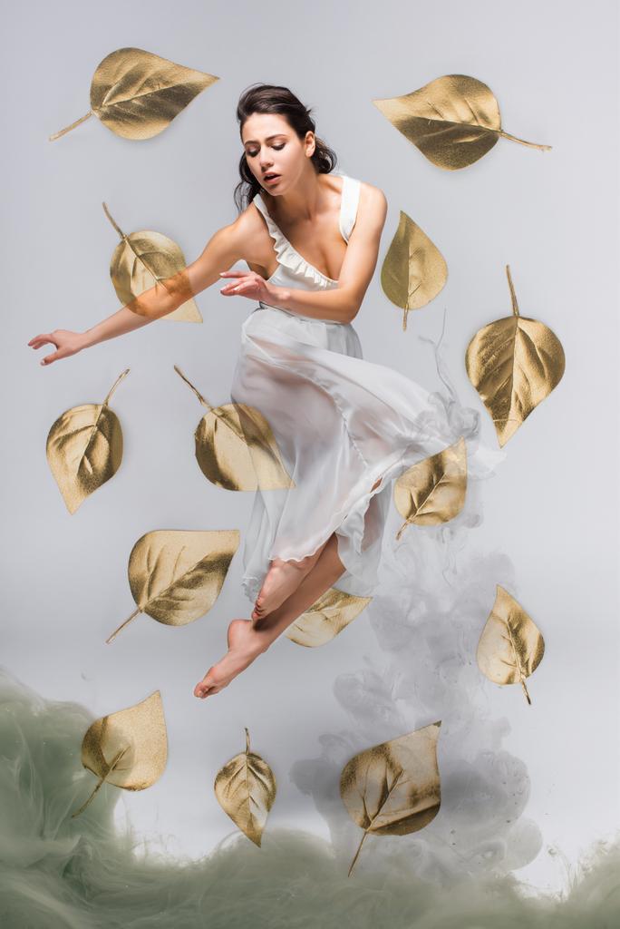 beautiful ballerina in white dress dancing surrounded by falling leaves near grey smoke on grey background - Photo, Image