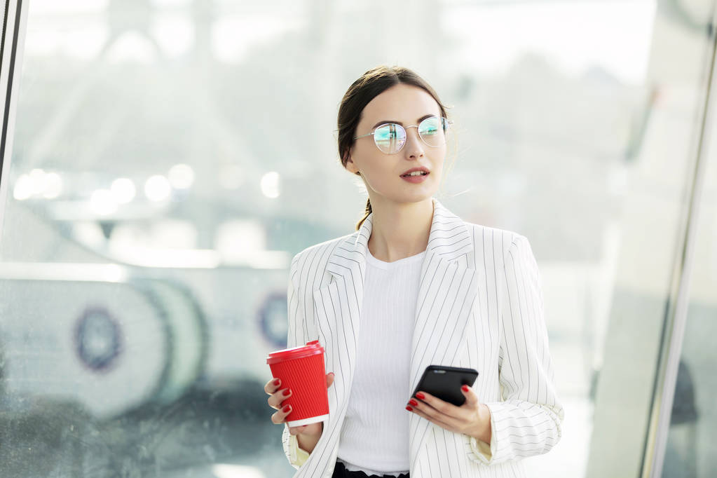 Rajattu kuva onnistuneen liiketoiminnan nainen yllään puku käyttäen moderni älypuhelin ja kahvin mennä ennen töihin aikaisin aamulla
 - Valokuva, kuva
