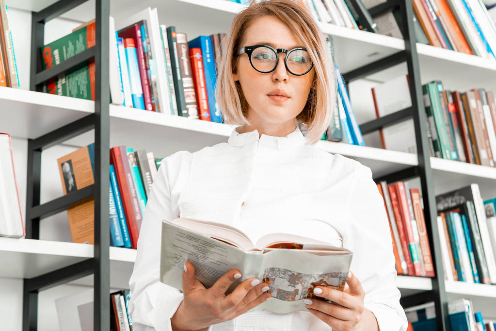 Шикарная привлекательная женщина в очках в формальной одежде читает книгу в библиотеке
. - Фото, изображение