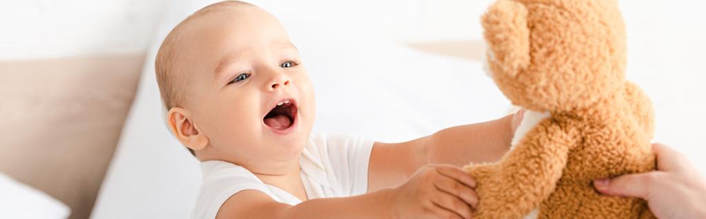 Foto panorámica de lindo niño pequeño sonriendo y levantando las manos para osito de peluche
 - Foto, imagen