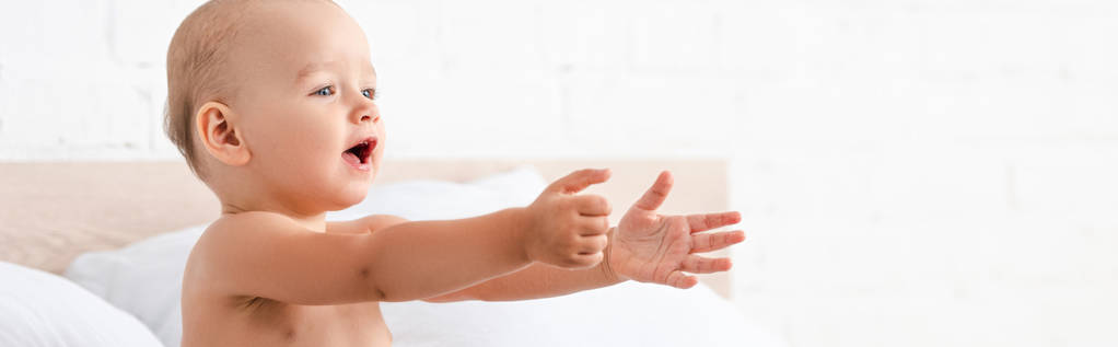 Panoramic shot of adorable little child opening mouth and raising hands forward in white room - Photo, Image