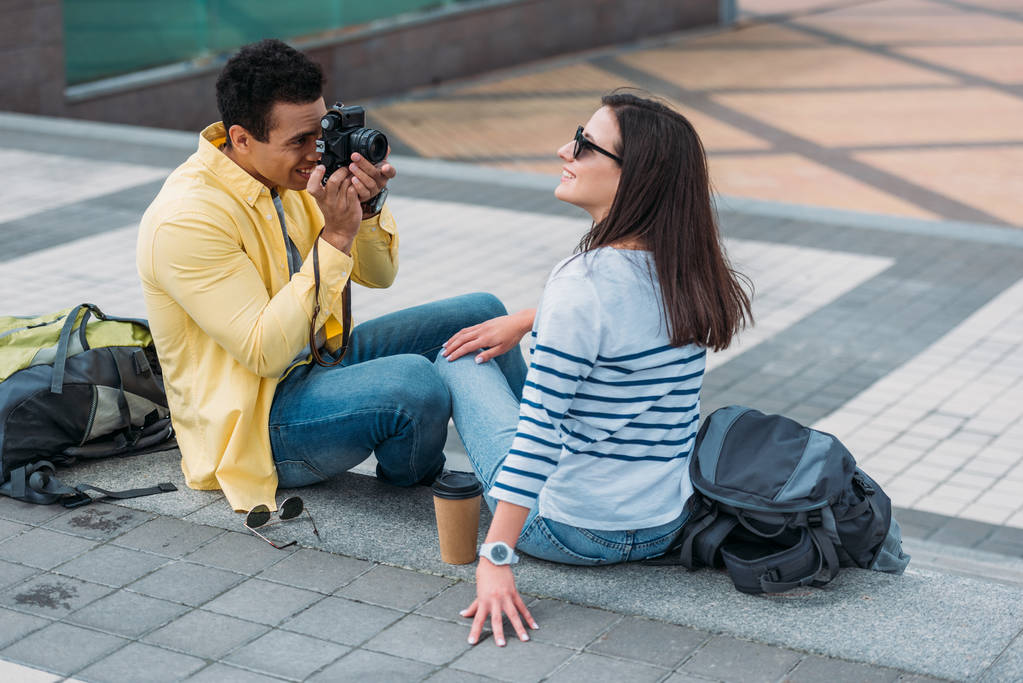 Bi-racial man sitting on stairs and taking photo of woman in sunglasses with backpack - Photo, Image