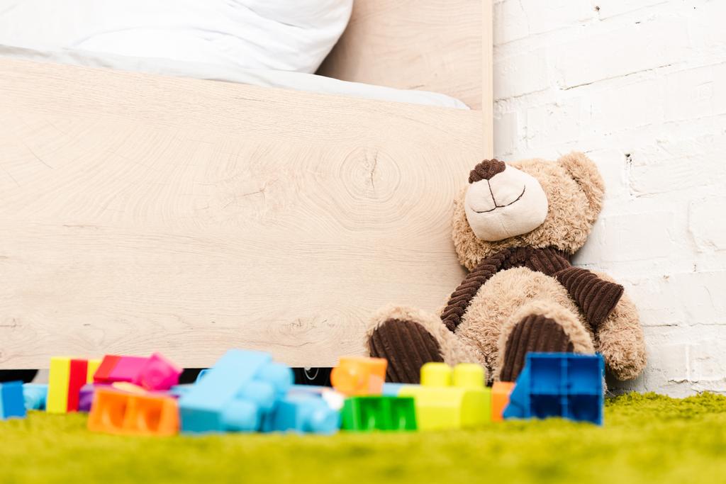 Βελούδινη αρκούδα και διάσπαρτα κομμάτια της κατασκευής σε πράσινο χαλί κοντά σε ξύλινο κρεβάτι - Φωτογραφία, εικόνα