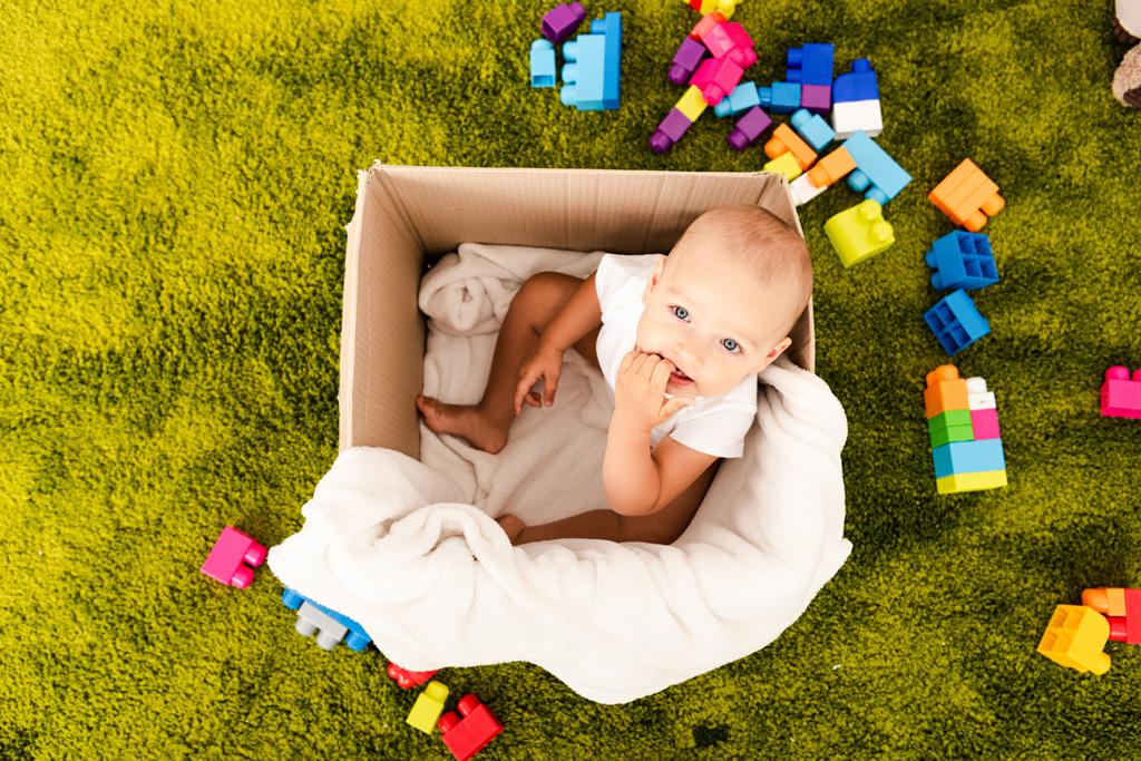 Top view of cute little child sitting in cardboard box with white blanket on green floor - Photo, Image