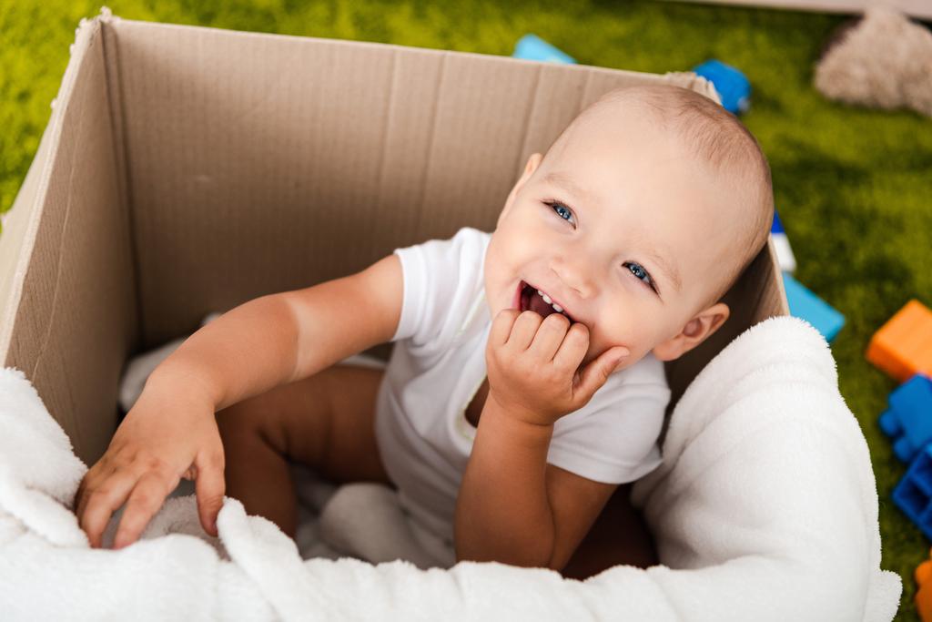 Criança bonito de olhos azuis sentado e sorrindo em caixa de papelão com cobertor
 - Foto, Imagem