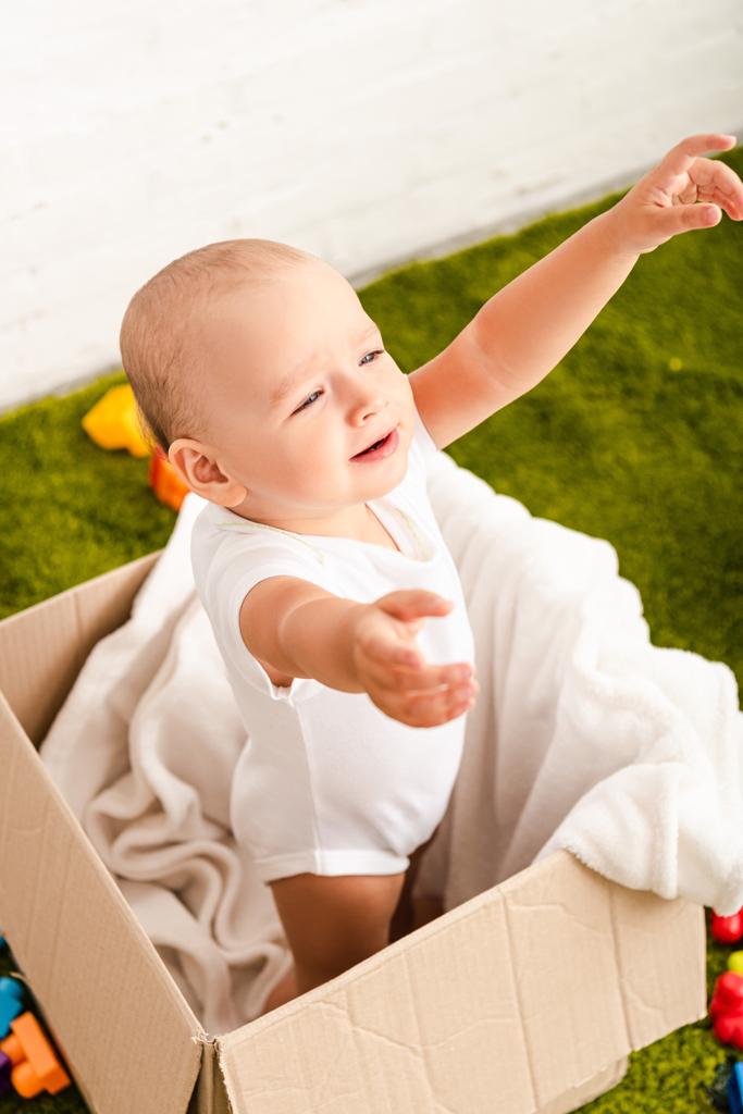Μικρό παιδί που στέκεται σε χάρτινο κουτί με λευκή κουβέρτα και σήκωσε τα χέρια - Φωτογραφία, εικόνα