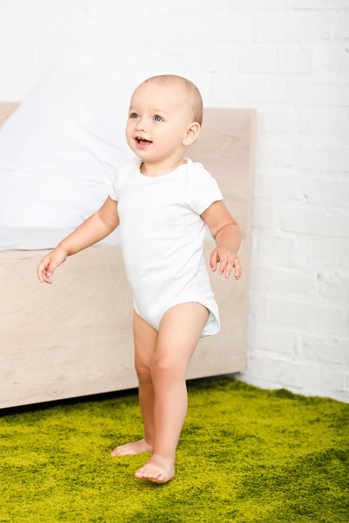 glückliches kleines Kind lächelt und geht durch helles Zimmer mit Bett und grünem Teppich - Foto, Bild