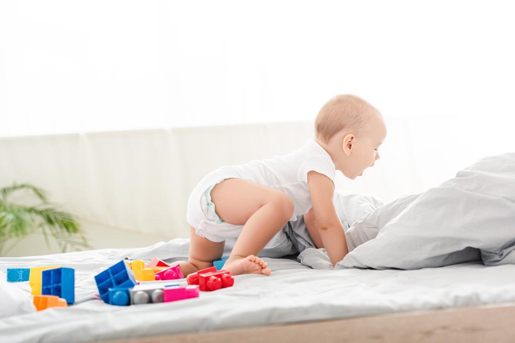 Śliczne boso Baby w białych ubraniach pełzające na łóżku wśród zabawek - Zdjęcie, obraz