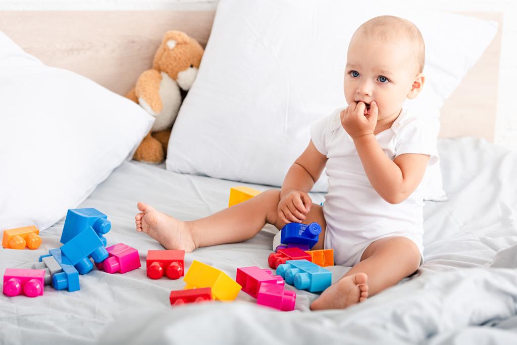 bebé descalzo con ropa blanca sentado en la cama con juguetes y tomando los dedos en la boca
 - Foto, imagen