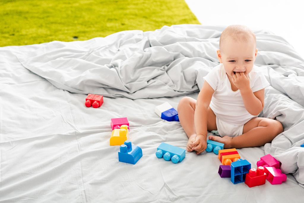 Hauska paljain jaloin vauva valkoisissa vaatteissa istuu sängyssä lähellä värikästä rakennetta ja ottaa sormet suuhunsa.
 - Valokuva, kuva