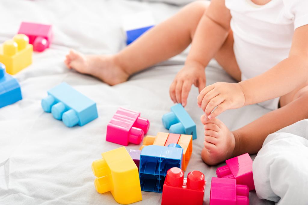Vista parcial del bebé descalzo en ropa blanca sentado en la cama y jugando con la construcción brillante
 - Foto, imagen