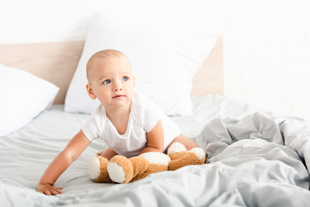 Carino bambino in abiti bianchi che tiene il suo orsacchiotto sul letto con cuscini
 - Foto, immagini