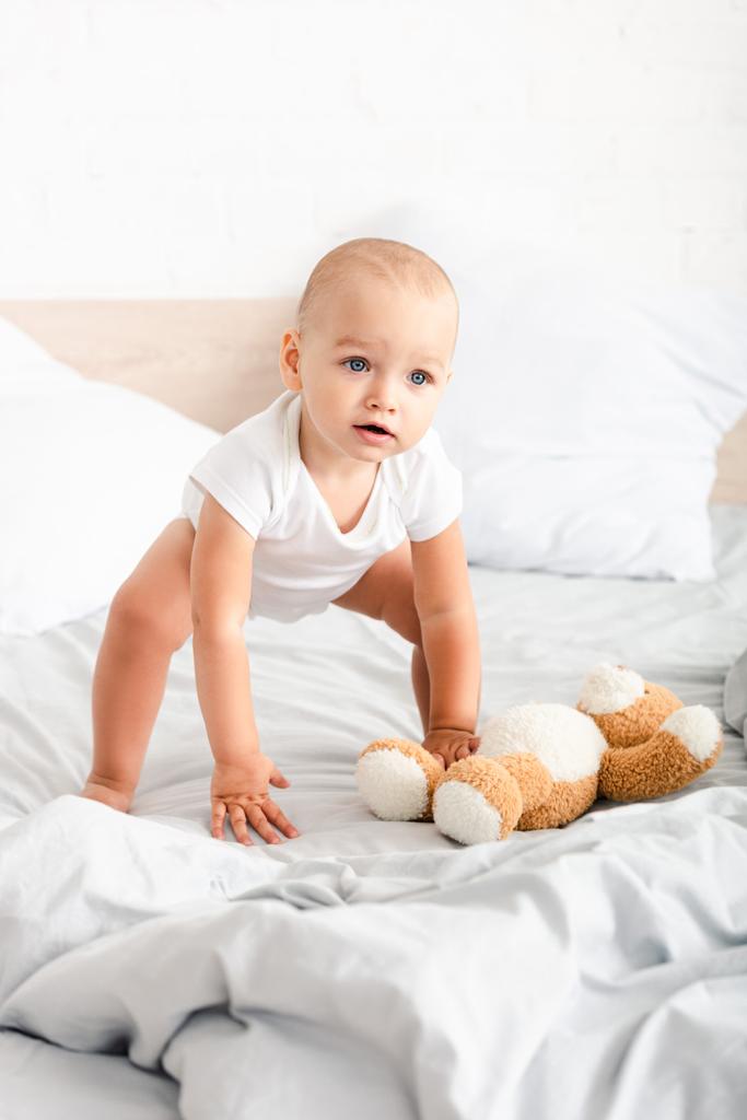 Χαριτωμένο παιδί σε λευκά ρούχα εκριζώματος αρκουδάκι του από το κρεβάτι - Φωτογραφία, εικόνα