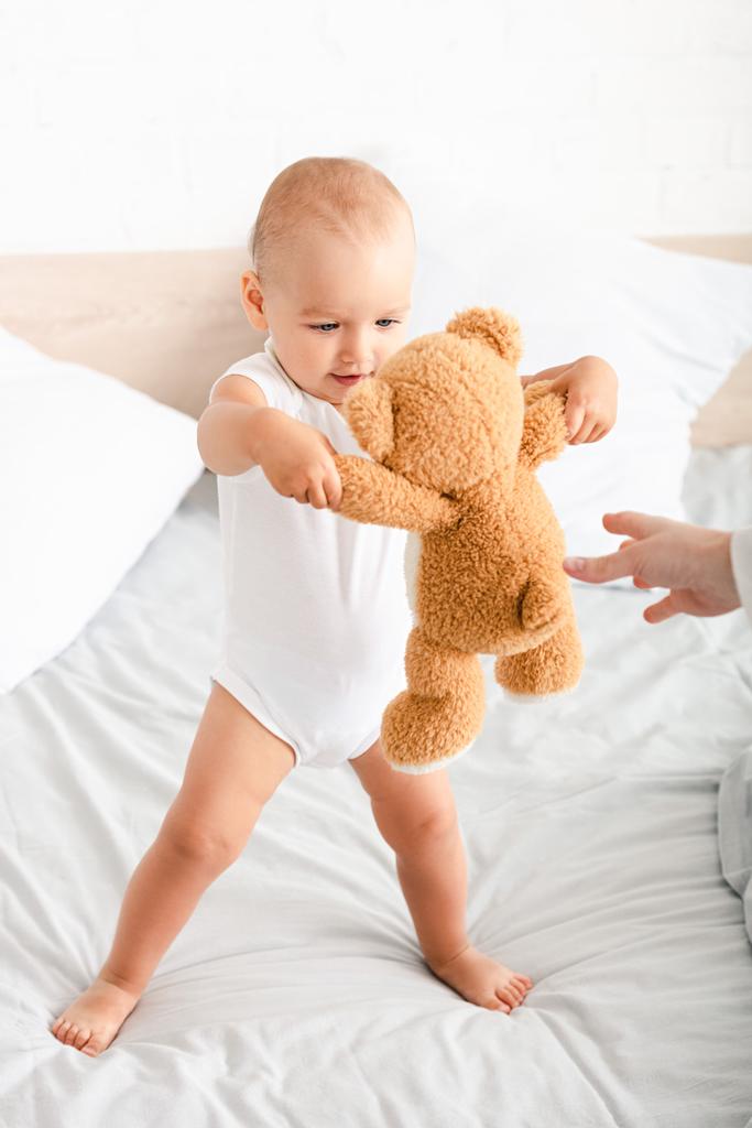 Χαριτωμένο ξυπόλυτο παιδί σε λευκά ρούχα παίζοντας με το αρκουδάκι του στο κρεβάτι - Φωτογραφία, εικόνα