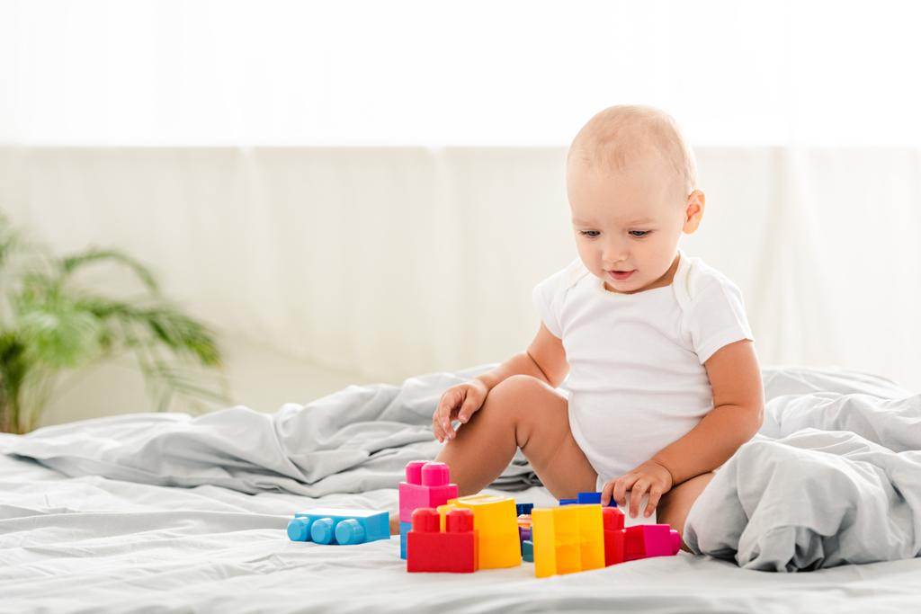 улыбающийся ребенок в белой одежде сидит на кровати и смотрит на игрушки
 - Фото, изображение