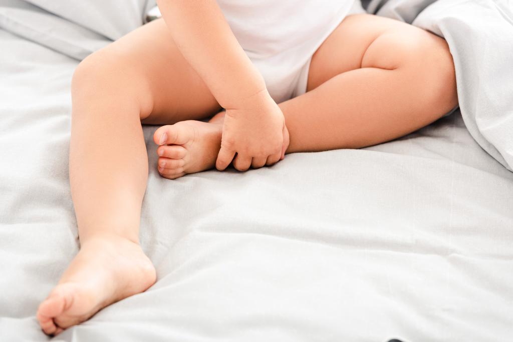 Περικομμένη θέα του χαριτωμένο μωρό κάθεται στο κρεβάτι και κρατώντας τα πόδια - Φωτογραφία, εικόνα