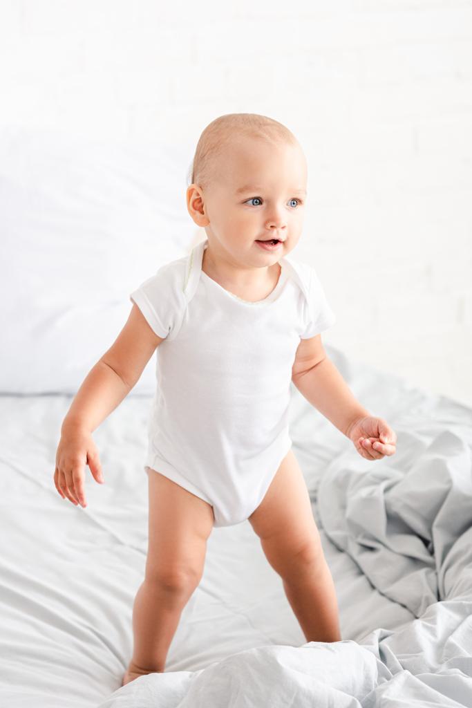 Śliczne małe dziecko w białych ubraniach stojących na łóżku, uśmiechając się i patrząc z dala - Zdjęcie, obraz
