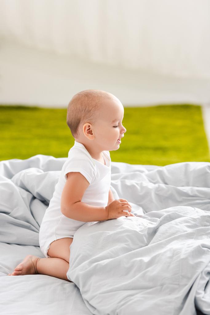 Boční pohled na malé dítě v bílých šatech, které sedí na posteli a dívá se dopředu - Fotografie, Obrázek