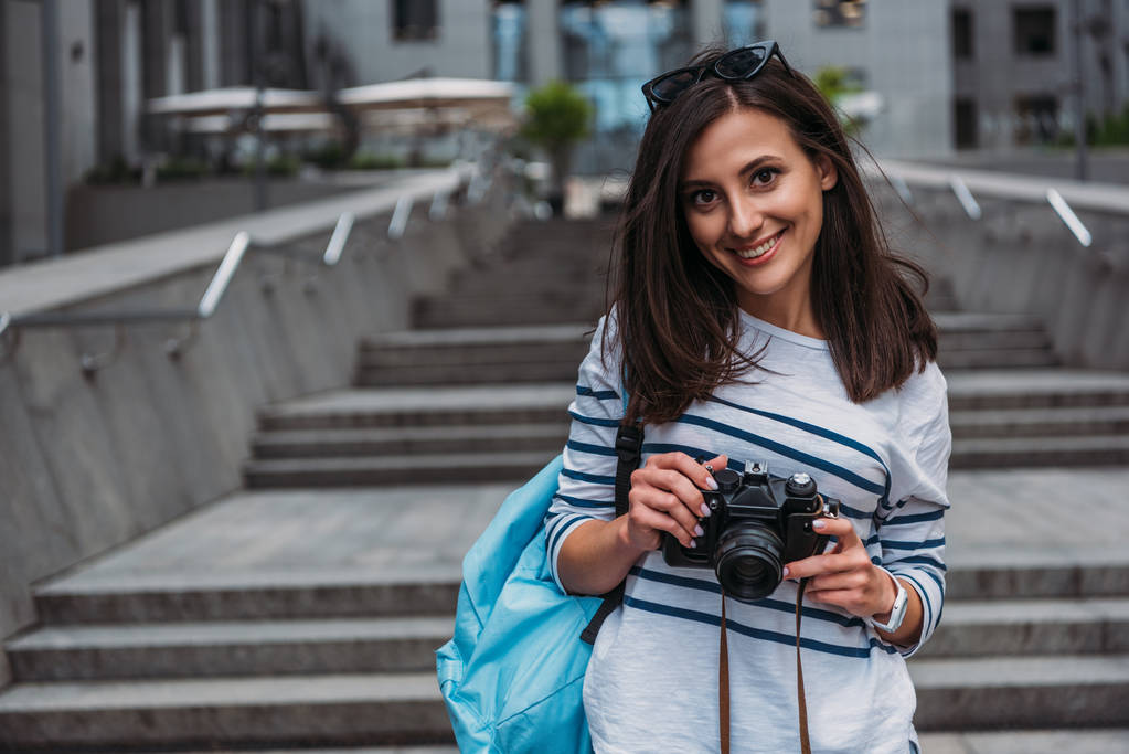 Mujer en casual con mochila sosteniendo cámara digital y sonriendo
 - Foto, Imagen