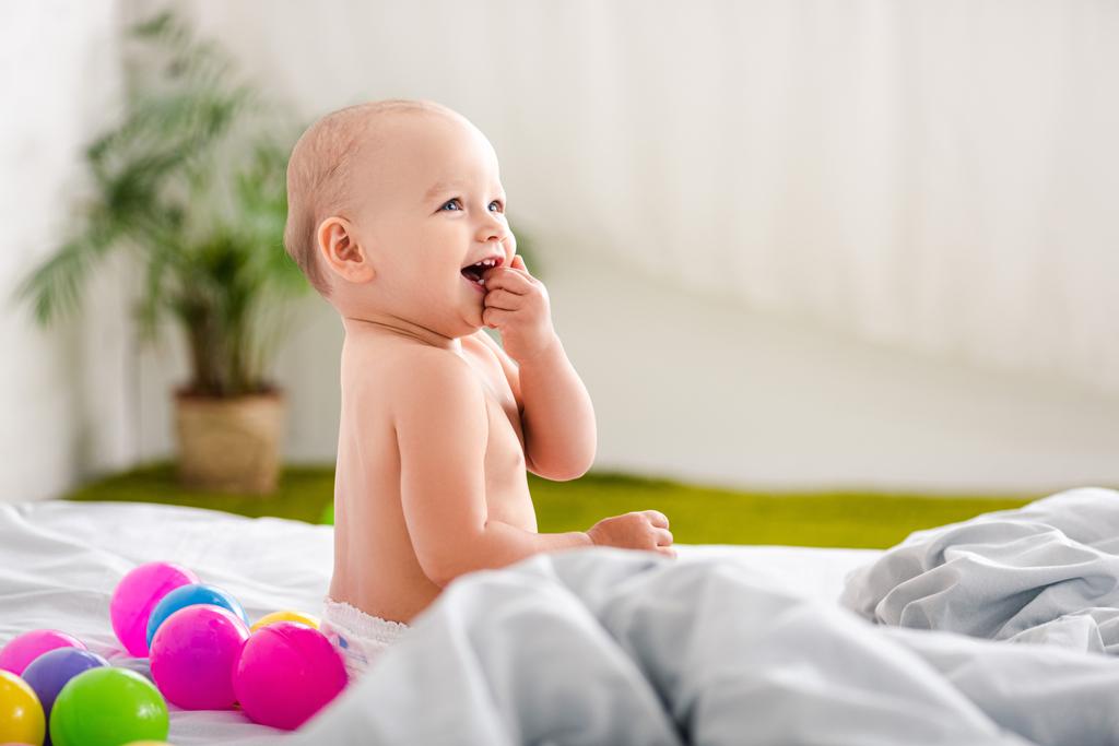 Criança bonita sentada na cama, sorrindo e tomando o dedo na boca
 - Foto, Imagem