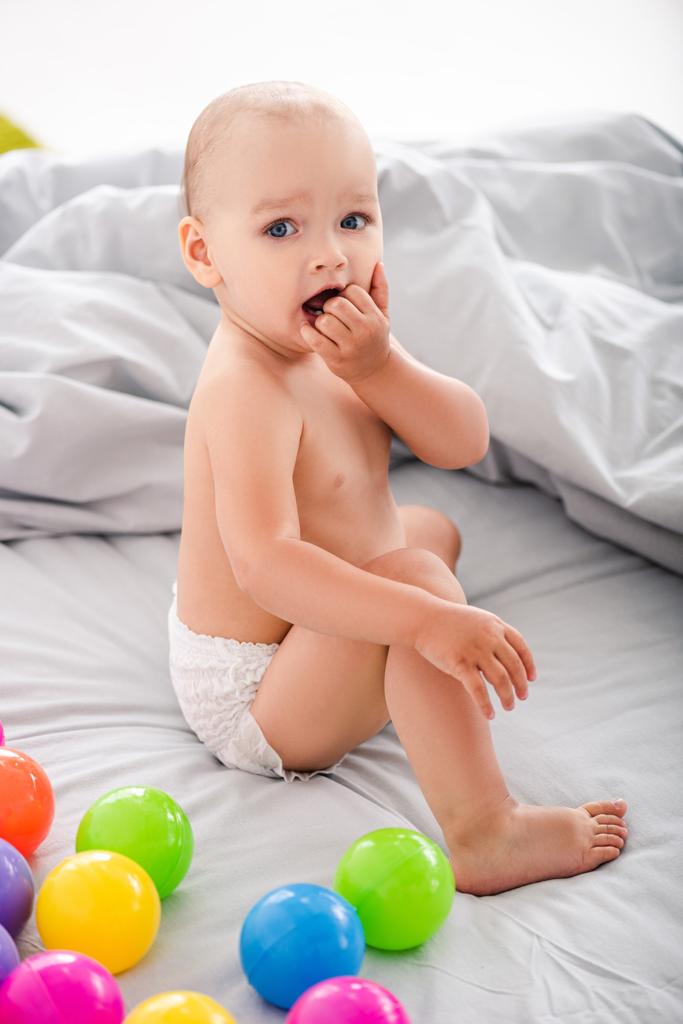 criança bonito sentado na cama entre bolas coloridas e levando os dedos na boca
 - Foto, Imagem