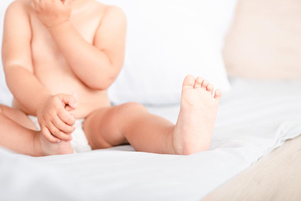 Vista parcial de criança pequena com dedos minúsculos e dedos dos pés sentados na cama branca
 - Foto, Imagem