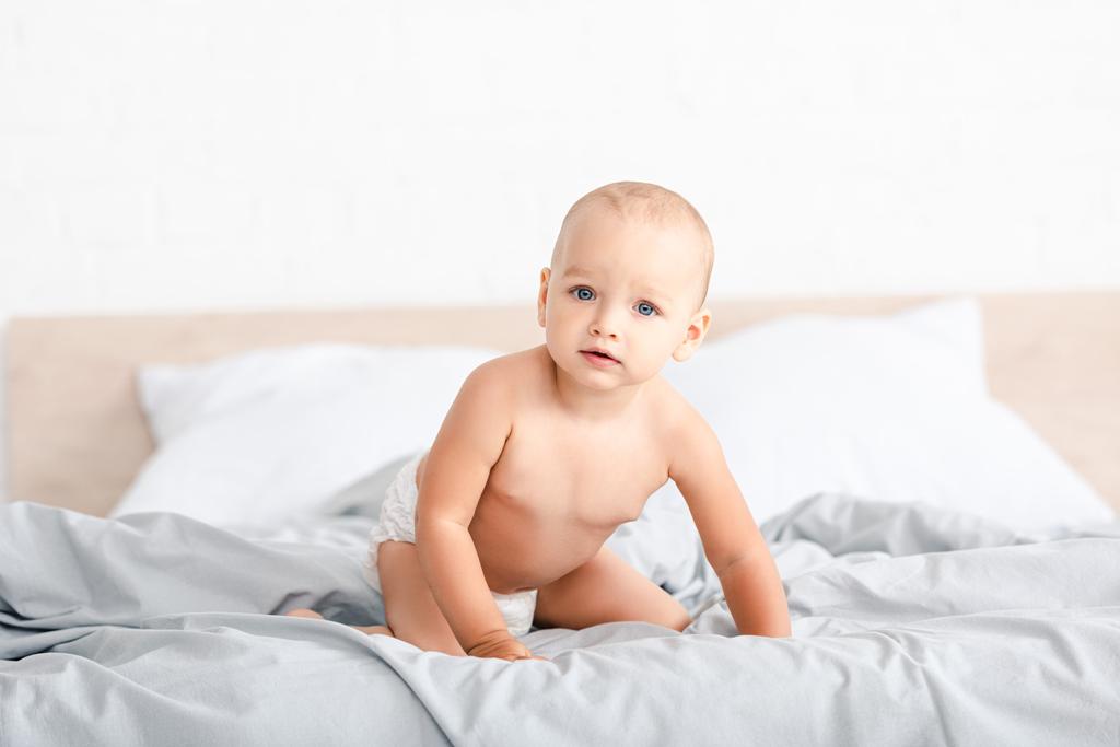 Симпатичный маленький ребенок сидит на белой кровати и смотрит в камеру
 - Фото, изображение