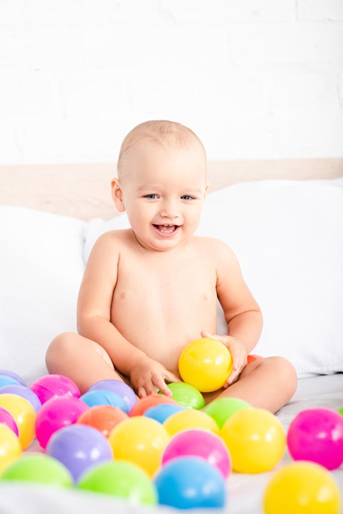 Χαριτωμένο μικρό μωρό κάθεται στο κρεβάτι, παίζοντας με έγχρωμες μπάλες και γέλιο - Φωτογραφία, εικόνα