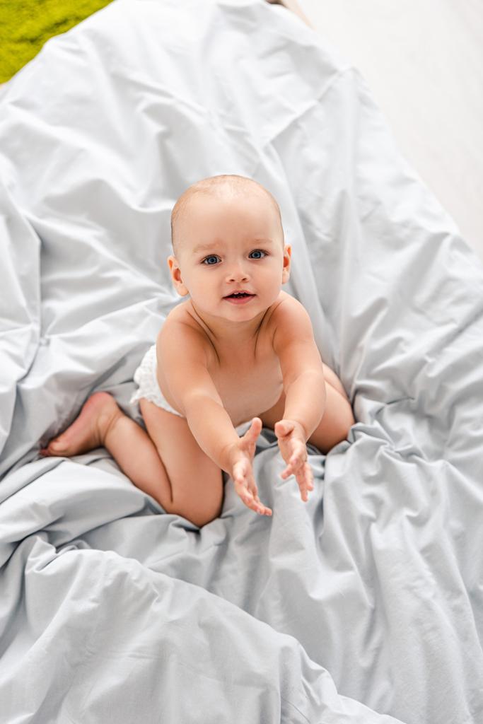 Χαριτωμένο μικρό μωρό σε πάνα κάθεται στο κρεβάτι και κοιτάζοντας την κάμερα - Φωτογραφία, εικόνα