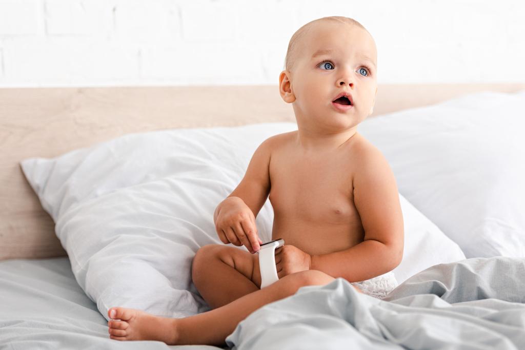 白いベッドに座って 手にスマートウォッチを保持しているかわいい赤ちゃん ロイヤリティフリー写真 画像素材