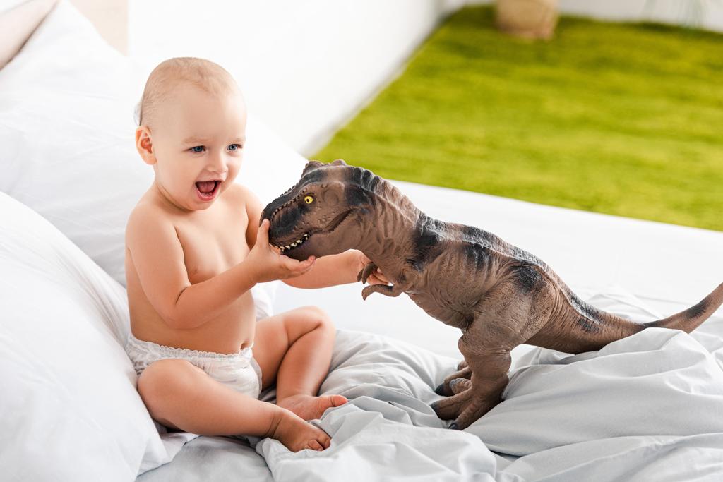 Cute little child smiling and playing with brown toy dinosaur on white bed - Photo, Image