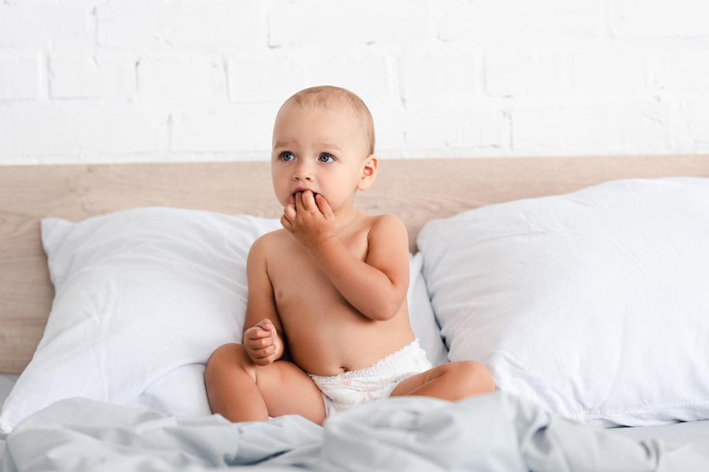 Очаровательный маленький ребенок сидит на кровати с подушками и берет пальцы в рот
 - Фото, изображение