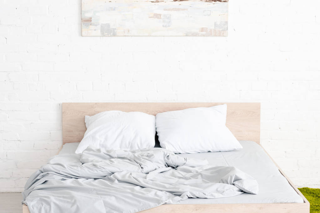 Ξύλινο κρεβάτι με δύο λευκά μαξιλάρια και τσαλακωμένη κουβέρτα στο φωτεινό δωμάτιο - Φωτογραφία, εικόνα