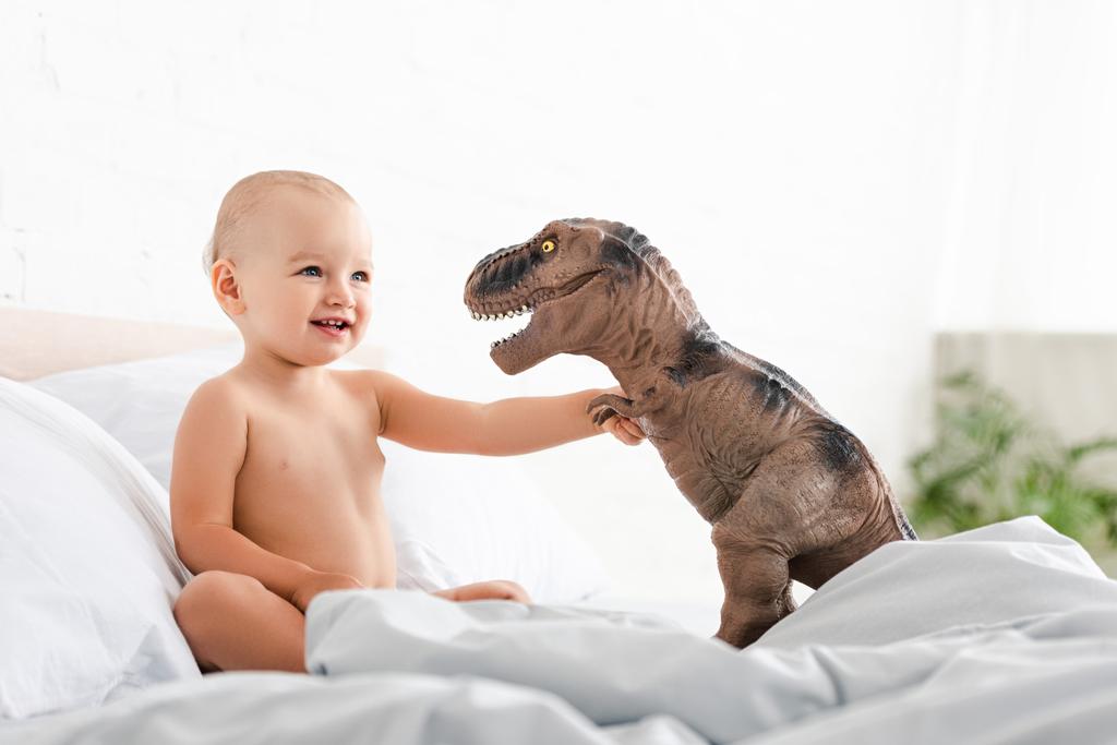 Lindo niño pequeño sentado en la cama, sonriendo y sosteniendo dinosaurio juguete marrón
  - Foto, imagen