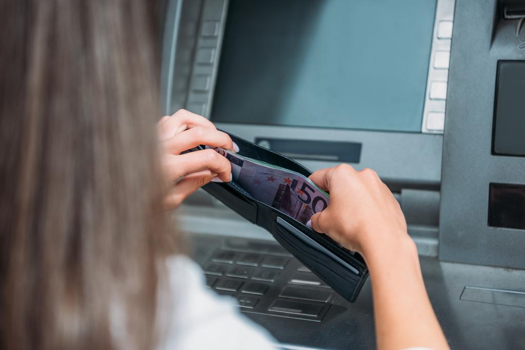 Ausgeschnittene Ansicht einer Frau, die in der Nähe eines Geldautomaten Geld in die Brieftasche steckt  - Foto, Bild