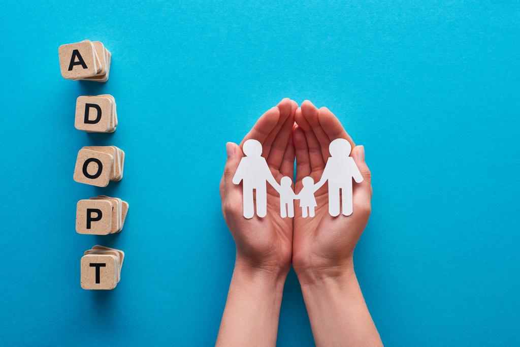 cropped view of woman holding paper cut gay family near blocks with adopt lettering on blue background - Photo, Image