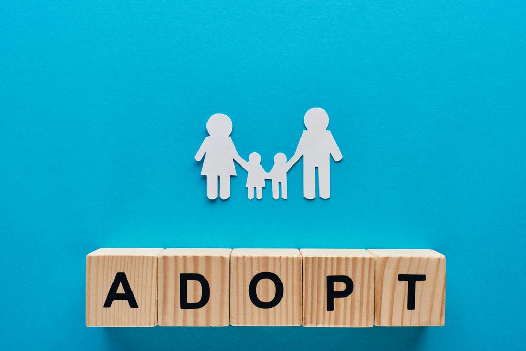 pohled na papír řezaných rodin držení rukou na modrém pozadí s adopci na dřevěných kostkách - Fotografie, Obrázek