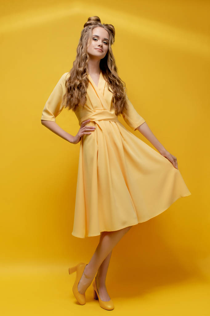Милая молодая девушка в жёлтом платье, в туфлях на каблуках на жёлтом фоне с стрижкой и кудрявыми длинными волосами. Желтая модная, стильная, молодежная одежда. Прически и макияж салона
. - Фото, изображение