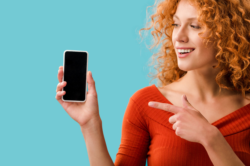 улыбающаяся рыжая девушка, указывающая на смартфон с пустым экраном, изолированным на синем
 - Фото, изображение