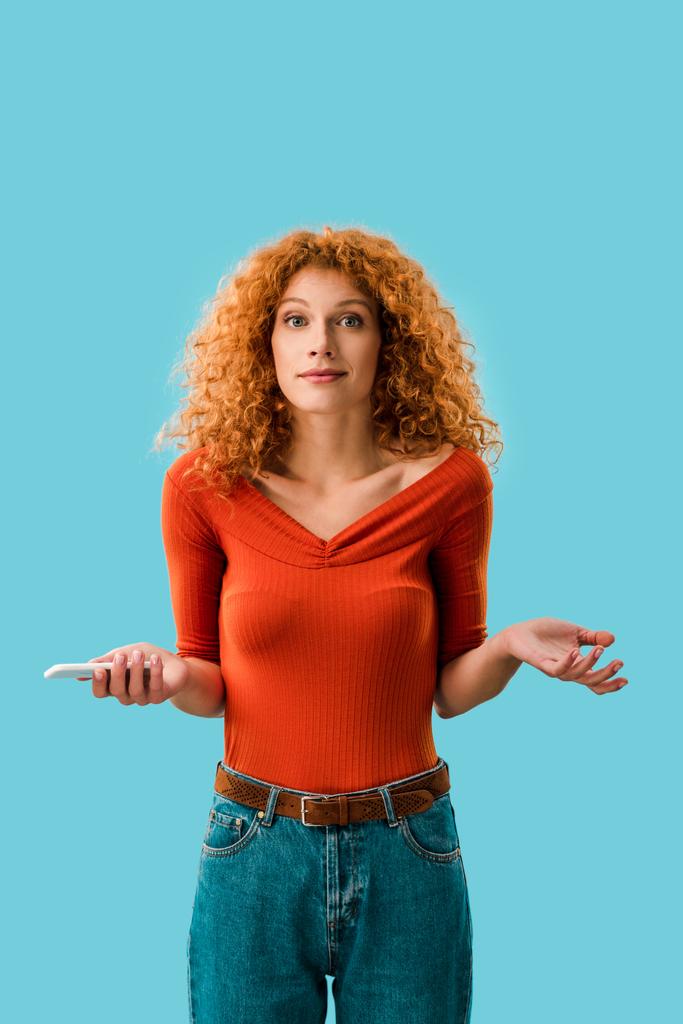 confused redhead woman holding smartphone isolated on blue - Photo, Image