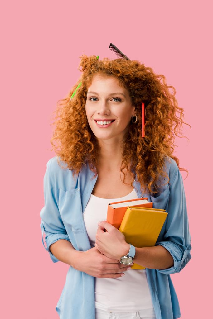 красивая улыбающаяся студентка с карандашами в волосах, держащая книги, изолированные на розовый
  - Фото, изображение