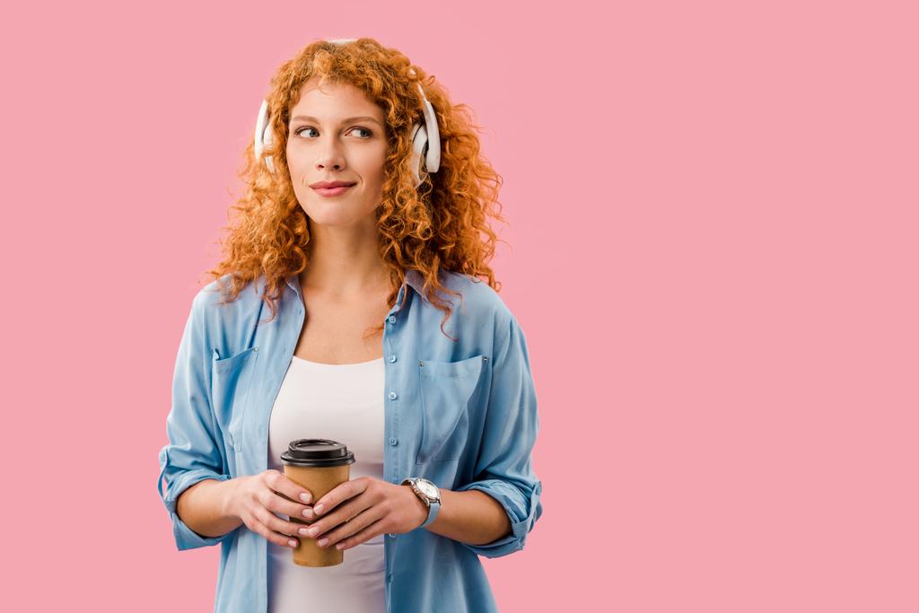 rudowłosy kobieta z kawą, aby przejść słuchanie muzyki w słuchawkach, izolowane na różowy - Zdjęcie, obraz