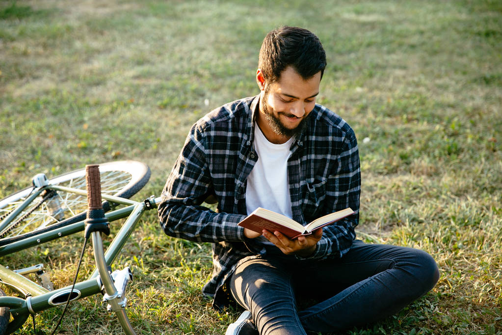 本を読んで、自転車の近くに座って、屋外で陽気な男. - 写真・画像