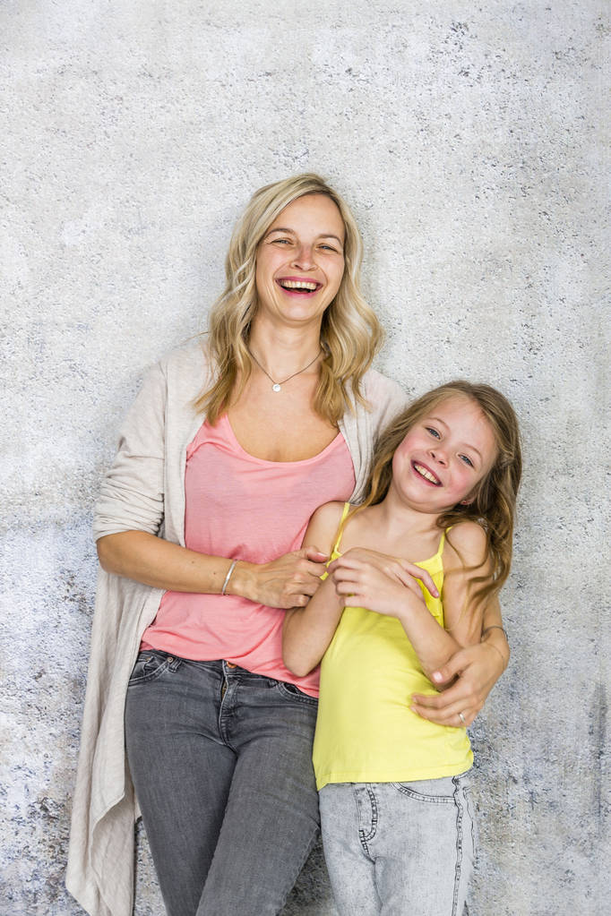 όμορφη ξανθιά νεαρή μητέρα που ποζάρει και αγκάλιασμα με την κόρη μπροστά από τσιμεντένιο τοίχο - Φωτογραφία, εικόνα