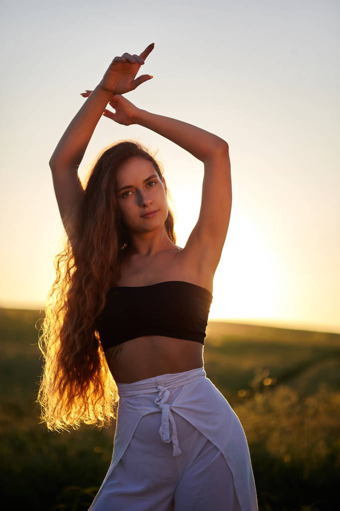 Portrait of beautiful young stylish girl in the sunlight at sunset. Posing.Fashion.Long haired,curly girl.She is cute.Beautiful woman is enjoying warm and sunlight. She is bathing in the sun - Photo, Image