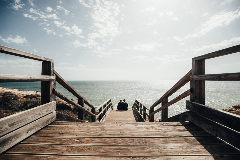 Pareja enamorada sentada en las escaleras cerca del océano. Atractiva pareja disfrutando de la vista al mar en el camino de madera. Hombre y mujer abrazándose y besándose en la playa de Portugal. Familia joven en un viaje
 - Foto, Imagen
