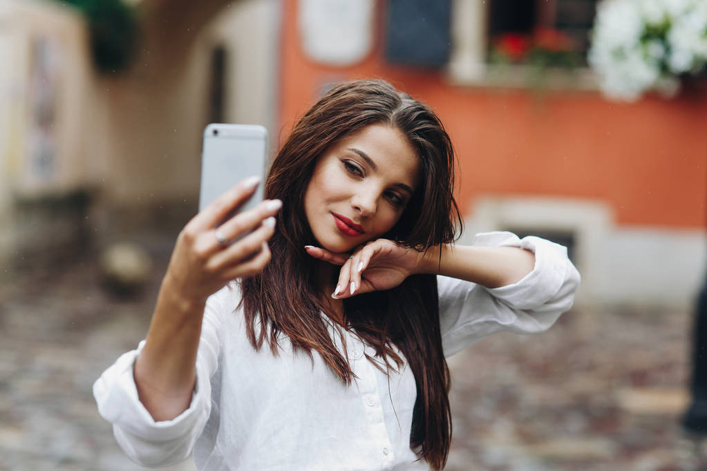 Горизонтальный портрет довольно счастливой молодой женщины в белой рубашке, делающей селфи или видео с фронтальной камерой на мобильном телефоне в городе
 - Фото, изображение