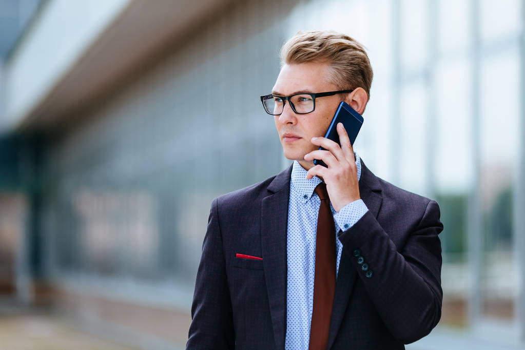 Nuori, komea, silmälasipäinen valkoihoinen liikemies puhuu matkapuhelimella toimistorakennuksen edessä. Mies toimitusjohtaja trendikkäissä vaatteissa älypuhelimella ulkona. Modernin teknologian käsite
 - Valokuva, kuva