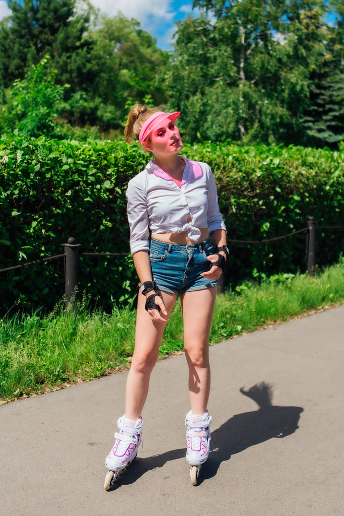 Retrato de una chica emocional en una visera de gorra rosa y guantes protectores para patines y patinetas montadas en patines en la carretera
. - Foto, imagen